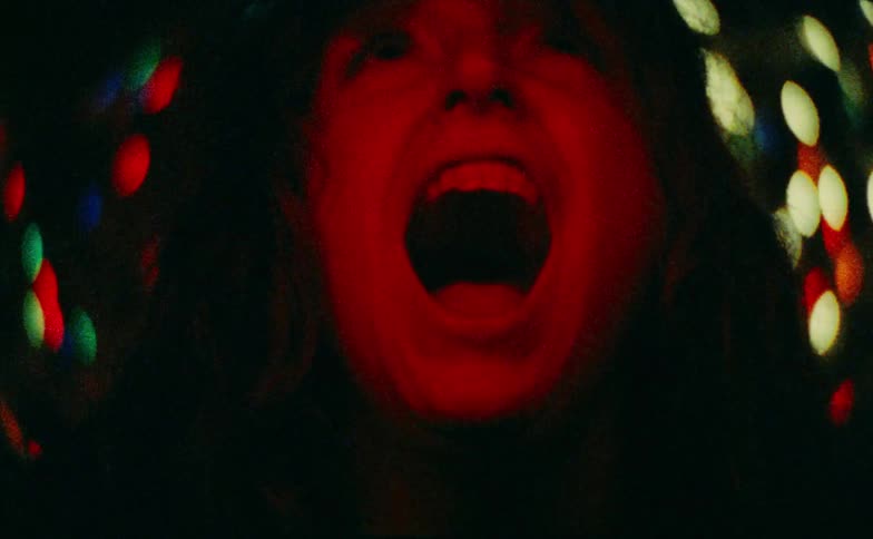 A young woman screams at the top of her voice while standing indoors in a dark room with a series of dim lights glowing behind her