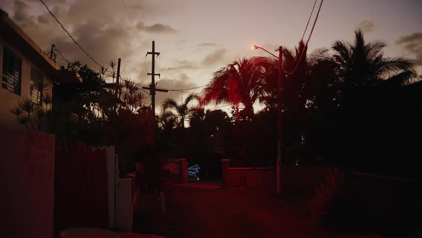 A street lamp glows over a quiet tropical neighborhood at dusk casting a warm orange light against the silhouettes of palm trees and utility lines under a cloudy evening sky