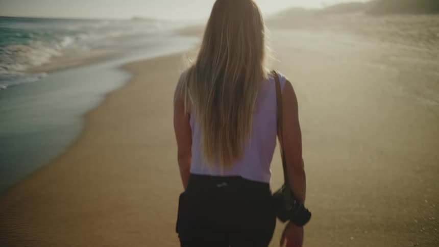 Blonde woman with a camera walking alone on the beach sand at dusk