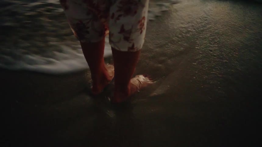A person stands on the sandy beach at midday as waves crash gently over their feet