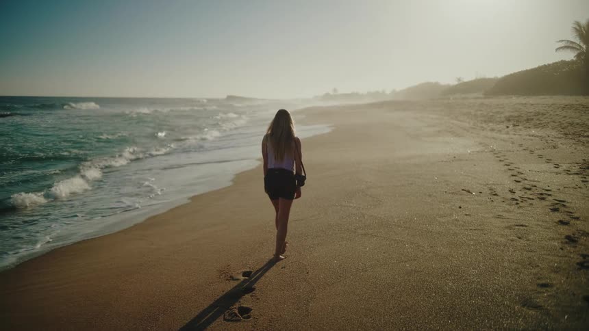 A woman walks alone along a sandy beach toward the bright sun leaving a trail of footprints behind her as ocean waves gently wash onto the shore