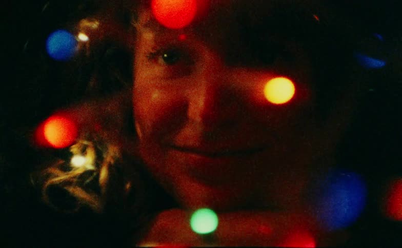 A young woman lies on her bed in a dark room holding a bundle of glowing christmas lights and smiling as she gazes at their warm glow