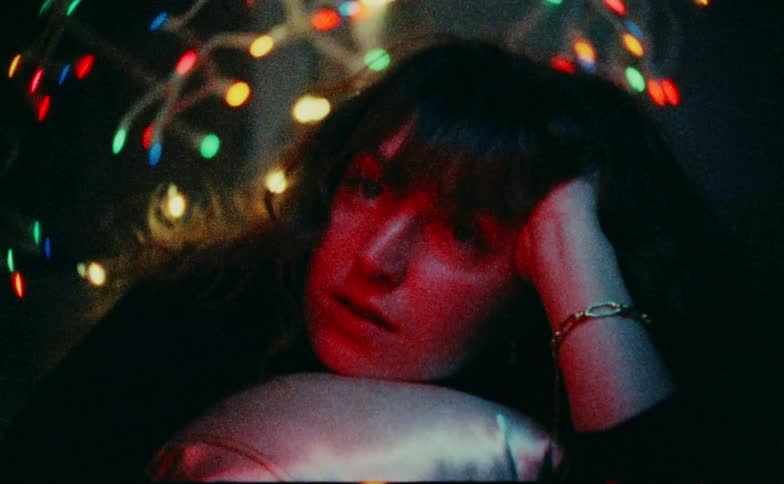 A pensive woman rests her head on a pillow as colored lights flash across her face in a dark grainy room