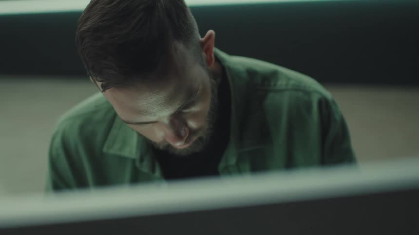 A bearded man in a green shirt sits at a desk and looks down with a focused expression while working behind a computer monitor in a dimly lit office environment