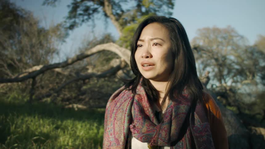 A woman standing outside on a sunny day in the woods shakes her head and wipes her forehead