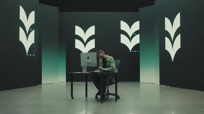 Man sitting at a table with a computer as he holds a pen and digital images move on the walls of the room