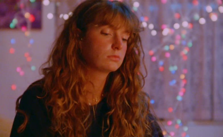 A young woman sits in a bedroom with her long hair falling over her shoulders surrounded by numerous glowing christmas lights shining behind her