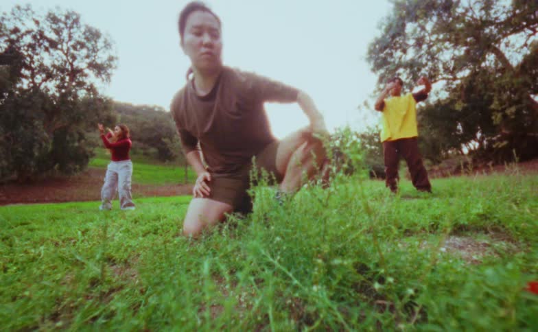 Three people dancing energetically on a grassy field surrounded by trees in daylight