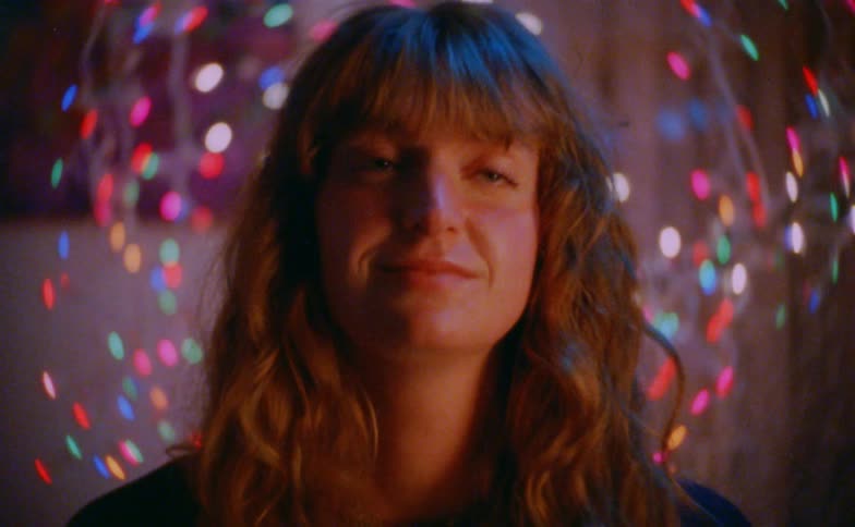 A young woman looks at the camera and slowly breaks into a gentle smile framed by a swirl of colorful out of focus lights