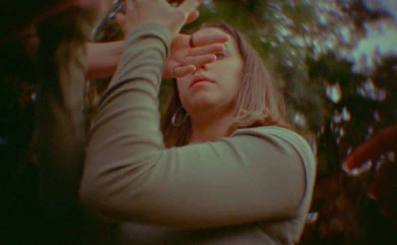 A woman with brown hair performs expressive fluid hand and arm movements while looking directly into the camera the scene is captured with a vintage slightly distorted lens effect against a backdrop of blurred trees