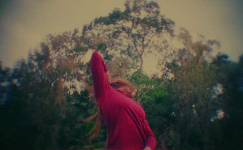 Person performs dance movements with arms extended in an outdoor setting surrounded by trees