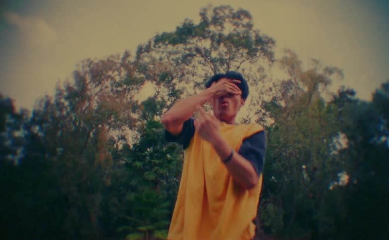 A man wearing a bright yellow vest over a dark shirt performs fluid and expressive dance movements with his hands while looking upward in an outdoor park setting with lush green trees