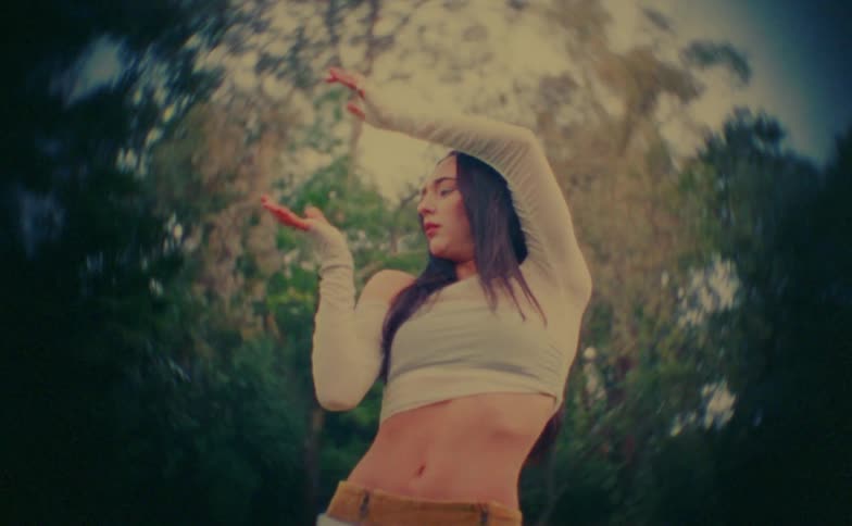 A young woman in a white crop top performs fluid expressive dance movements with her arms while standing in a lush wooded outdoor environment with a distorted lens effect