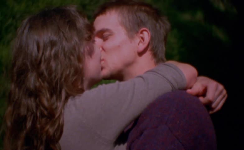 A man and a woman stand outdoors in a close embrace smiling warmly at each other before sharing a gentle kiss against a softfocus background of green trees and natural sunlight