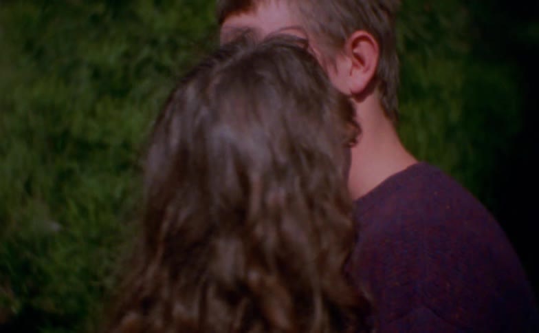 Adult people stand face to face very close together in an outdoor setting with greenery in the background