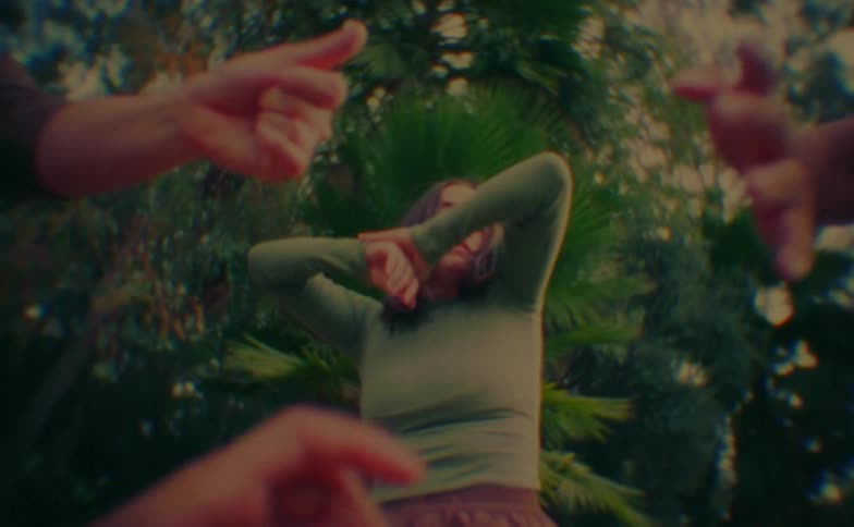 A young woman performs fluid dance movements in a garden while framed by several hands reaching into the shot from a lowangle perspective against a backdrop of lush palm trees and greenery