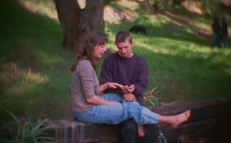A couple sits side by side in a public park at midday watching content on a mobile device while a dog wanders through the grass near a nearby tree behind them