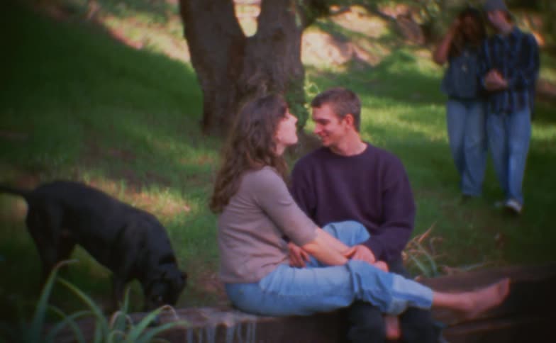 The black dog is playing behind the couple sitting on the groundthere is another couple walking towards the couple sitting with the dog