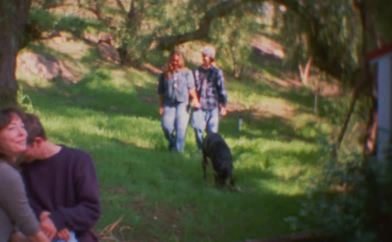 People sitting on the grass watch others walking with a dog in a wooded area