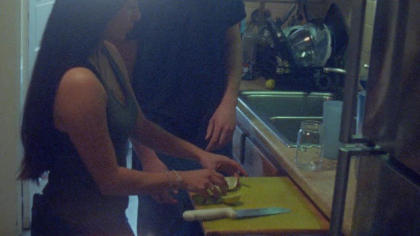 Person slicing green fruit on pullout board while other holds cup and gestures in kitchen
