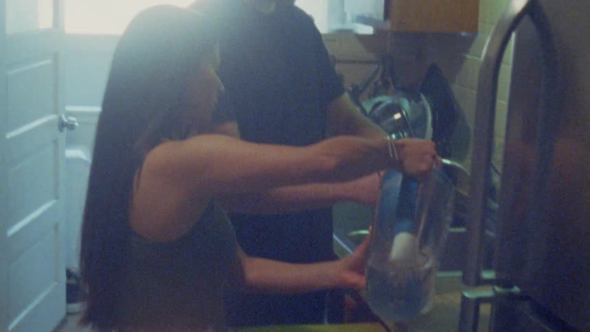 A woman in a grey tank top pours water from a blue filtered pitcher into a glass while a man stands beside her in a dimly lit kitchen featuring a cutting board and a refrigerator
