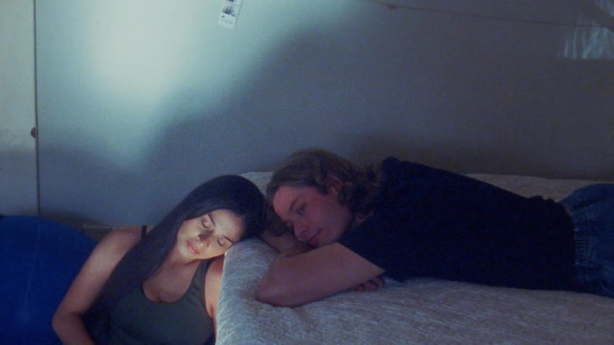 The woman sits next to the bed where a man is laying