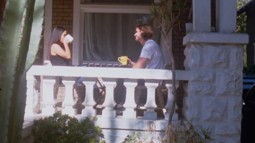 Young adult man and young adult woman toasting with coffee cups and sipping from them on a porch in front of a house outdoors during day time