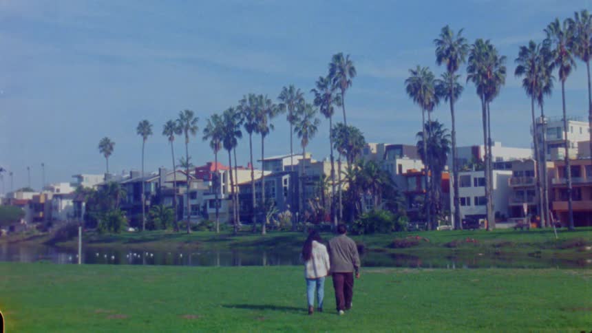Couple walking side by side on grassy field with modern buildings and palm trees in the background