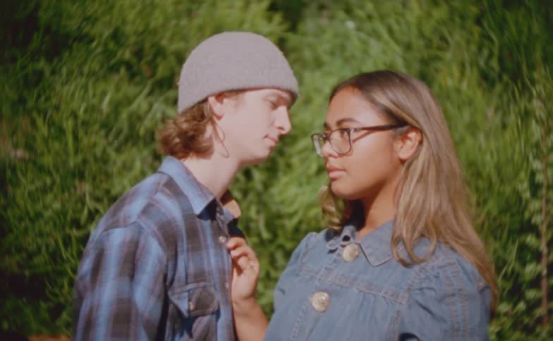 A male teen kissing his female partner on her cheeks while standing outdoors during daytime