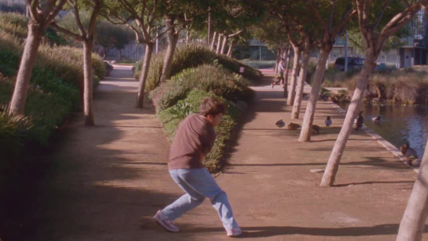 A young boy in a brown shirt and blue jeans performs expressive dance movements on a dirt path lined with trees next to a pond