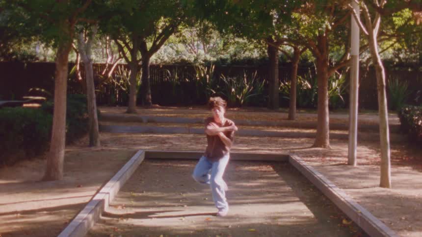 A young man performs a series of fluid rhythmic dance moves inside a woodenbordered park court surrounded by trees