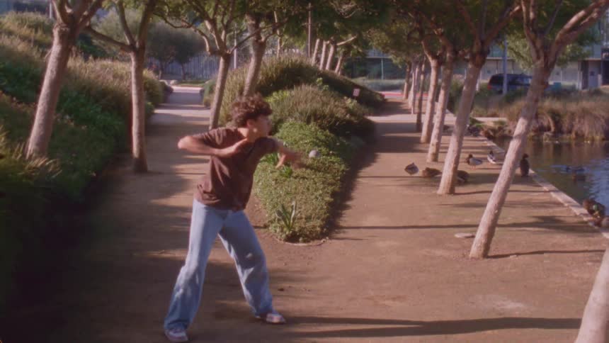 A young man performs expressive dance moves on a dirt path in a sunny park lined with trees ducks and a nearby pond