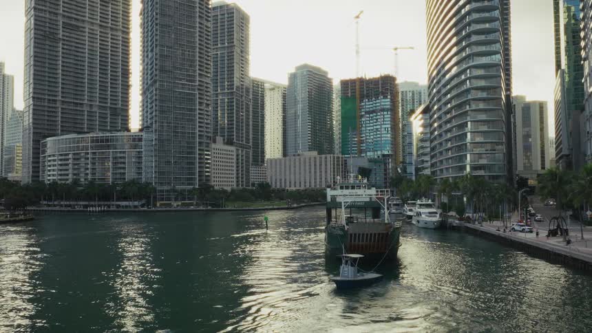 A cargo boat travels along a city river lined with modern high rise buildings during daylight