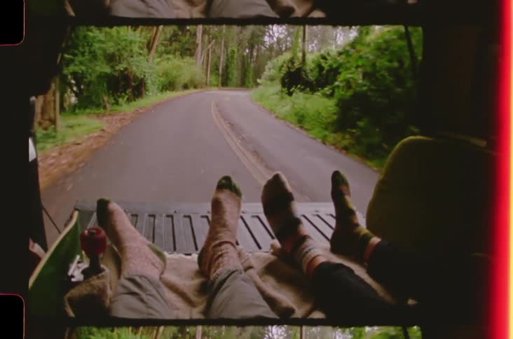 Two people rest their feet in the back of a moving vehicle that travels down a winding road through a dense green forest