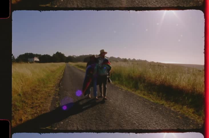 Couple walks arm in arm along a rural road surrounded by fields under direct sunlight