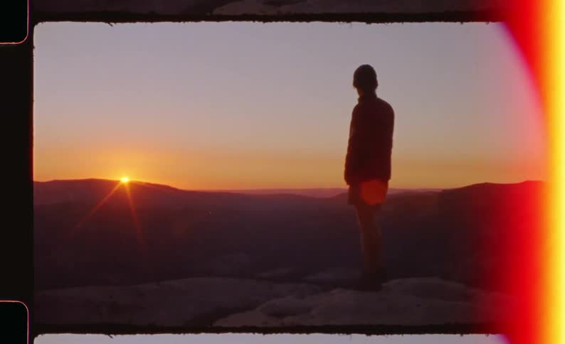 A person stands at the top of a mountain gazing toward the setting sun as warm light spreads across the horizon