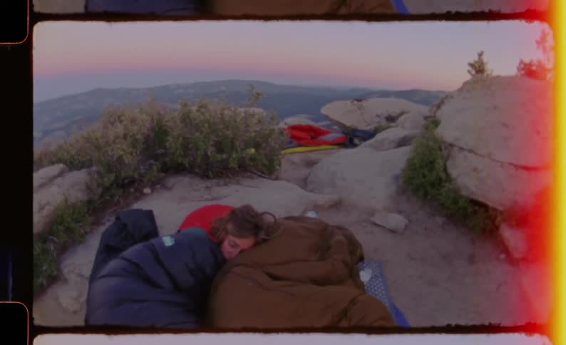 A woman sleeps inside a blue sleeping bag on a rocky mountain peak at dawn while surrounded by camping gear and a panoramic view of distant valleys under a clear twilight sky