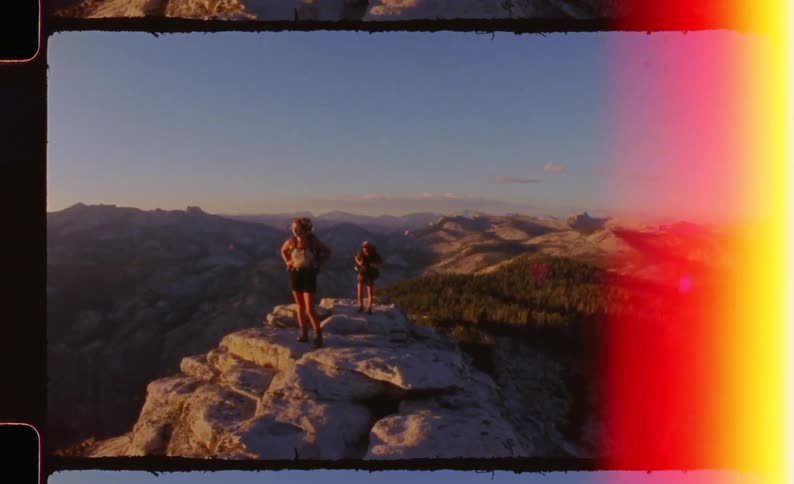 Two women stand atop a rugged lightcolored stone summit overlooking a vast expanse of mountain ridges and valleys with a blue sky