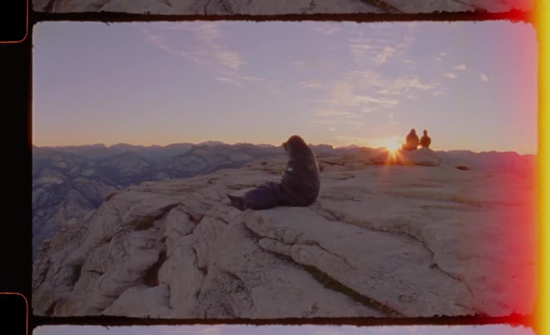 Three people sit atop a mountain at sunset watching the natural landscape in the warm fading light two sit together at a distance while a woman sits alone near the edge gazing out over the view