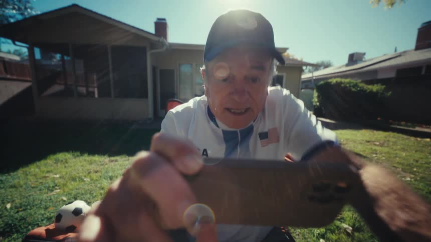 An older man wearing a usa hat and shirt sits in a lawn chair and reacts with surprise while holding a smartphone toward the viewer in a sunny backyard
