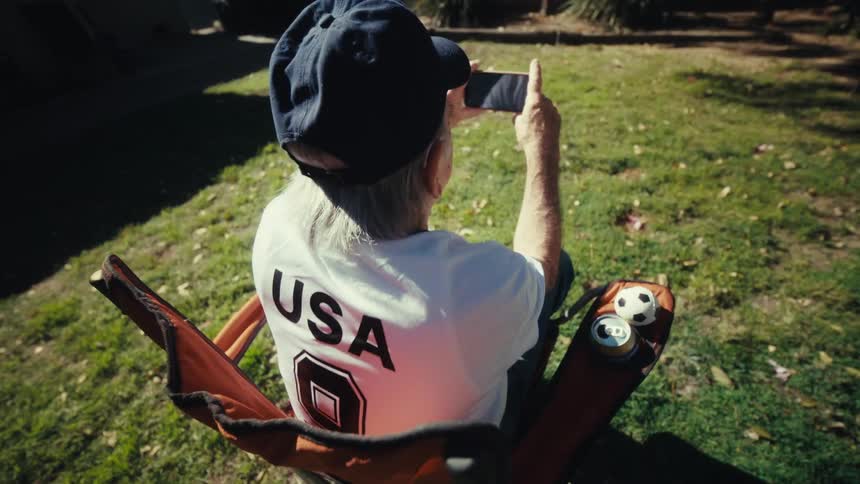 An elderly man is shown from the back sitting on a chair in a yard cheering while watching a smartphone screen