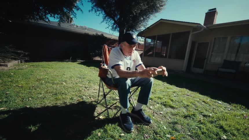 An elderly man sits in a folding chair in a sunny backyard holding a smartphone horizontally with both handshe appears focused on the screen while leaning forward slightly surrounded by a green lawn and residential buildings