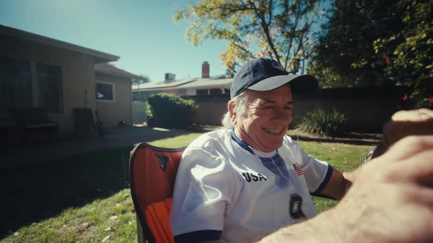 An elderly man wearing a usa soccer jersey sits in a folding chair in a sunny backyard while holding a smartphone horizontallyhe reacts with excitement by raising his fist and smiling widely as he looks at the device under the bright sun
