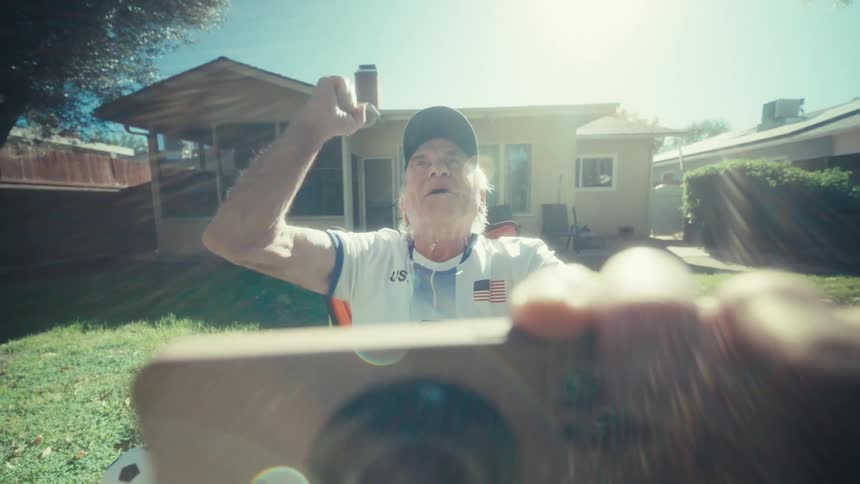 An elderly man sits in a folding chair in a grassy backyard while holding a smartphone horizontally with both hands he wears a white jersey blue cap and sneakers as he focuses on the phone screen under bright sunlight