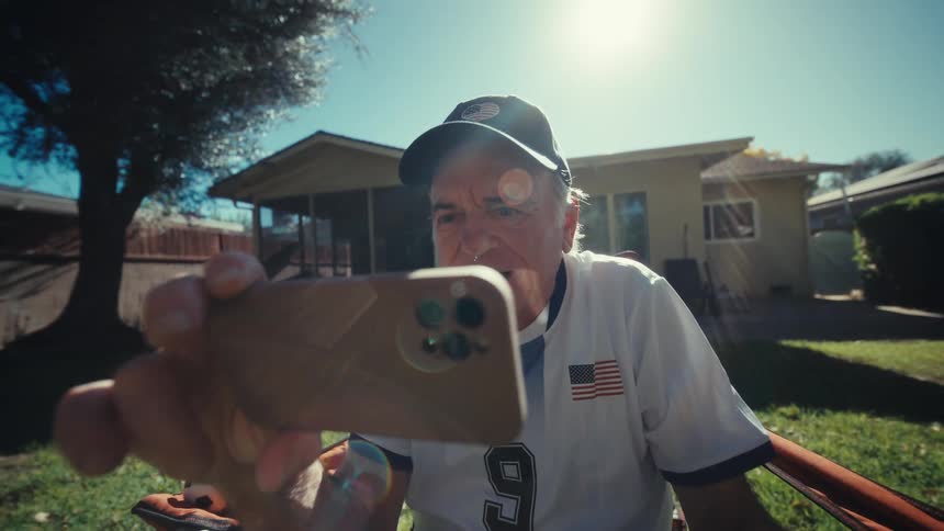An elderly man cheers while watching sports on his smartphone in a backyard during the day