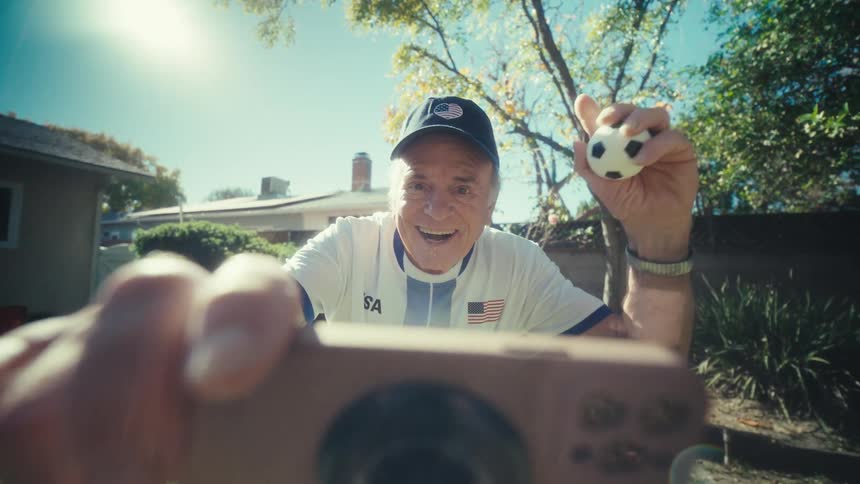 An elderly man in a usa soccer jersey celebrates enthusiastically while being recorded on a smartphone in a sunny backyard he raises a small soccer ball and pumps his fist in the air with a wide smile of excitement
