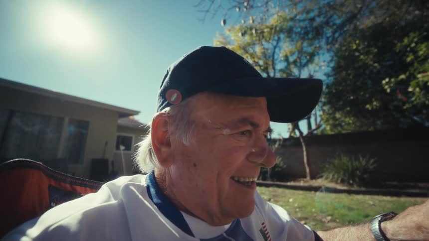 An elderly man sits in a lawn chair in a sunny backyard holding a smartphone to watch something while reacting with intense excitement laughter and celebratory gestures directed toward the camera lens