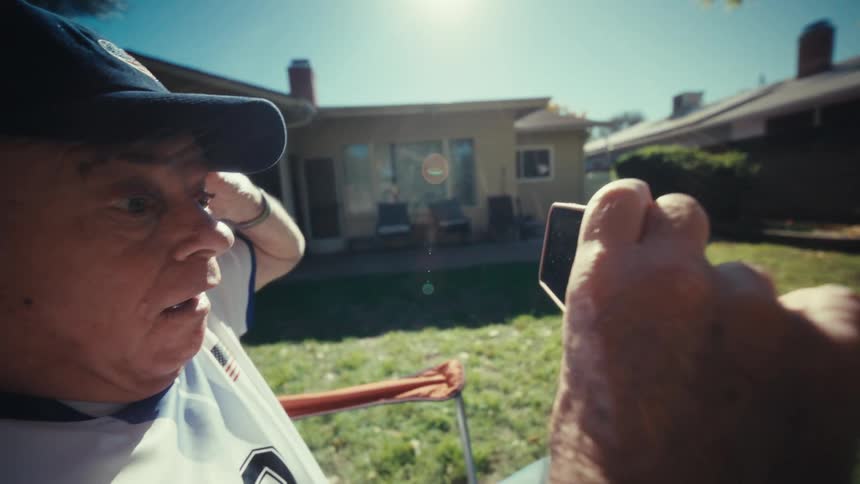An elderly man wearing a usa jersey and a baseball cap sits in an outdoor folding chair while watching something on his smartphonehe appears excited and gestures with his hands while looking at the screen in a sunny backyard setting