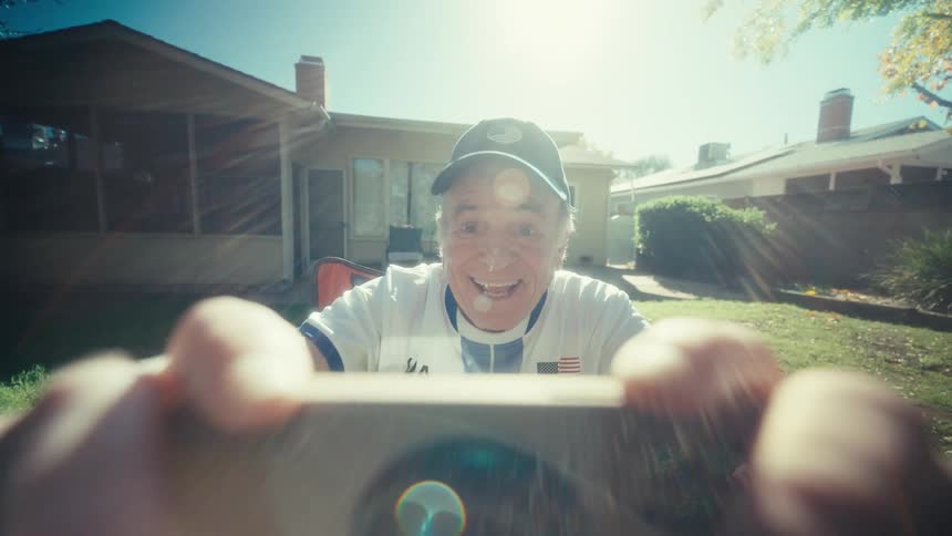 An excited elderly man cheers while holding a smartphone in a sunny suburban backyard