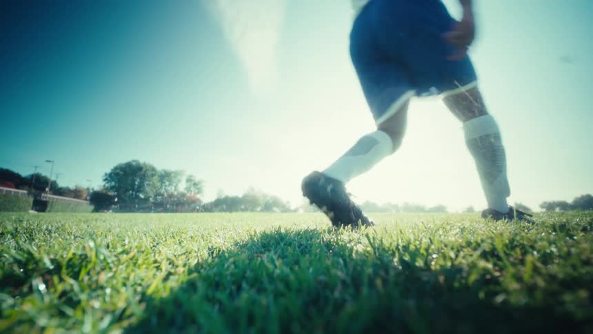 A football player kicks a soccer ball on a sunny grass field wearing blue trunks and a white jersey as he strikes the ball with power and focus
